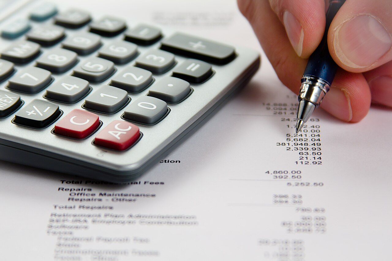 Mão com caneta confere os números de um relatório financeiro ao lado de uma calculadora